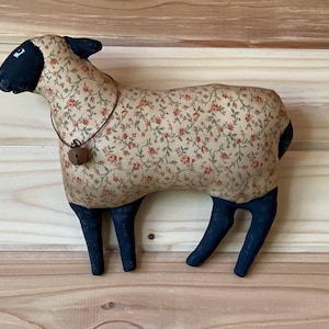 May include: A stuffed sheep figurine with a beige floral pattern body, black legs, head, and tail. It has a bell around its neck. The sheep is a decorative item, likely for home decor or a child's room.