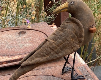 May include: A handcrafted, brown fabric bird with a yellow beak and metal legs. The bird has a heart-shaped charm around its neck and is perched on a rusty metal surface. The bird's wings are textured, and the overall design has a rustic, vintage aesthetic.