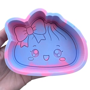 May include: A silicone mold featuring a swirled pink and blue color pattern. The interior design depicts a rounded character face with large eyes, a smiling mouth, and a bow positioned on the upper left side of the head.