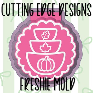 May include: A pink cookie cutter mold featuring a scalloped circular frame with a stacked design of three bowls. The top bowl has an oak leaf, the middle has a maple leaf, and the bottom has a pumpkin. The text CUTTING EDGE DESIGNS and FRESHIE MOLD is visible.