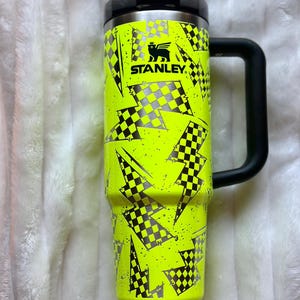 K&ouml;nnte beinhalten: Eine gelbe Stanley-Reise-Thermoskanne mit schwarzem Griff. Die Kanne hat ein Schachbrettmuster und ein Blitz-Muster.