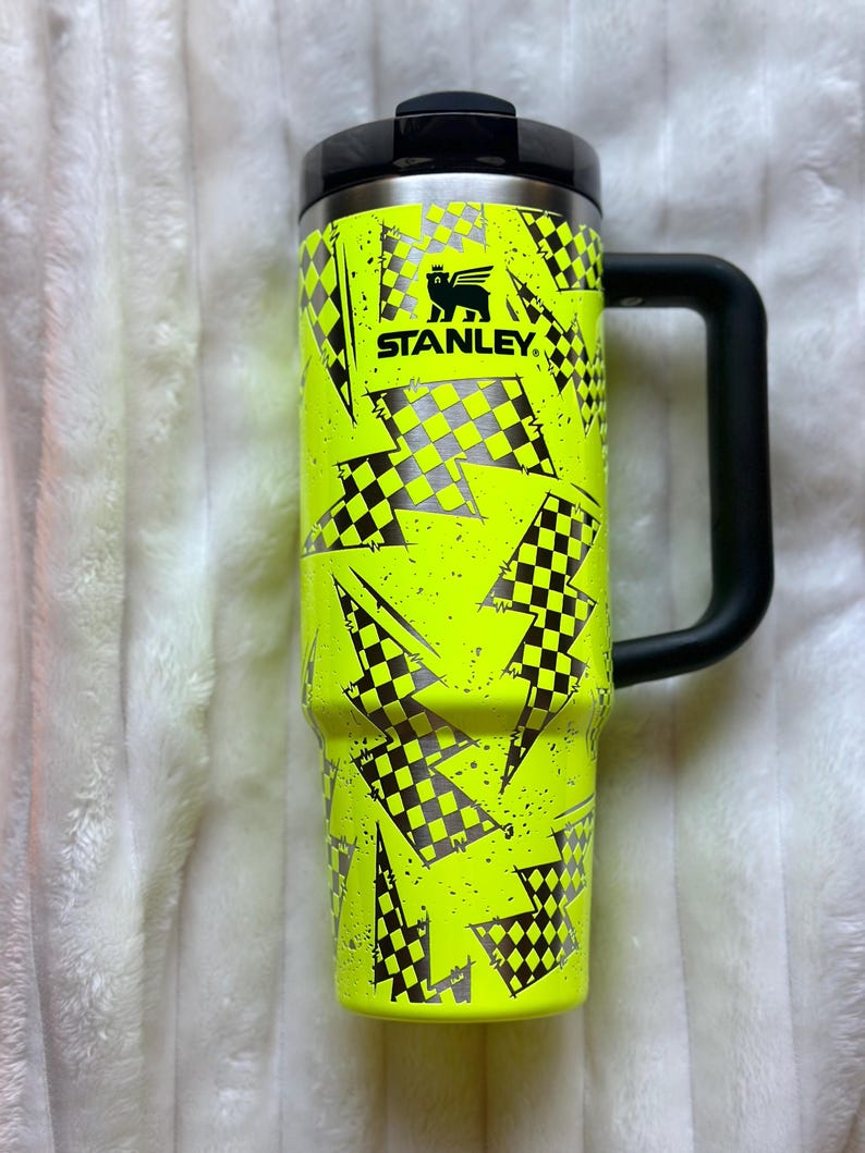 K&ouml;nnte beinhalten: Eine gelbe Stanley-Reise-Thermoskanne mit schwarzem Griff. Die Kanne hat ein Schachbrettmuster und ein Blitz-Muster.