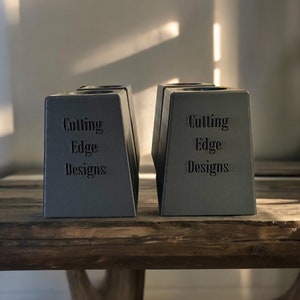 May include: Two gray, triangular-shaped candle holders with the text "Cutting Edge Designs" on the front.