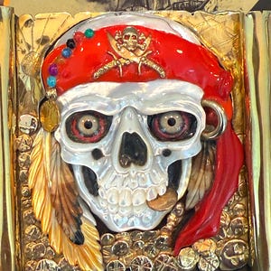 Könnte beinhalten: Detaillierte, goldfarbene Gürtelschnalle mit Piratenmotiv. Die Schnalle zeigt einen Totenkopf mit rotem Bandana, goldenen Ohrringen und einem goldenen Emblem. Der Totenkopf hat detaillierte Augen und eine Goldmünze im Mund. Die Schnalle ist von goldenen Akzenten umgeben.