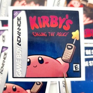 Peut inclure: Un autocollant repr&eacute;sentant une cartouche de jeu vid&eacute;o pour Game Boy Advance. L'autocollant pr&eacute;sente un personnage rose tenant un t&eacute;l&eacute;phone avec le texte "KIRBY'S CALLING THE POLICE" en lettres rouges.