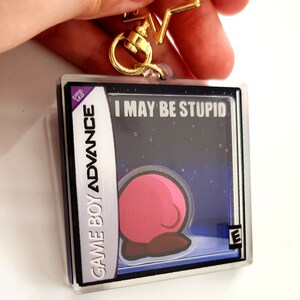 Peut inclure: Un porte-cl&eacute;s en plastique transparent en forme de cartouche Game Boy Advance avec un personnage rose sur un fond bleu fonc&eacute;. Le texte sur la cartouche indique "I MAY BE STUPID" et "GAME BOY ADVANCE". La classification "E" est visible dans le coin inf&eacute;rieur droit.