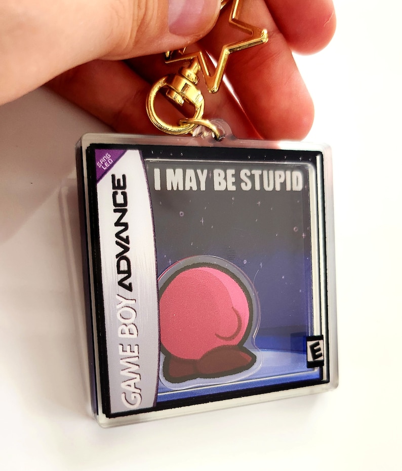 Peut inclure: Un porte-cl&eacute;s en plastique transparent en forme de cartouche Game Boy Advance avec un personnage rose sur un fond bleu fonc&eacute;. Le texte sur la cartouche indique "I MAY BE STUPID" et "GAME BOY ADVANCE". La classification "E" est visible dans le coin inf&eacute;rieur droit.