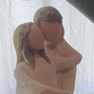 Puede incluir: Una figura tallada que representa a una pareja abrazándose. Las figuras son de tonos neutros, con la mujer con un vestido largo. El hombre está detrás de ella, con los brazos alrededor de ella. El fondo es blanco y texturizado.
