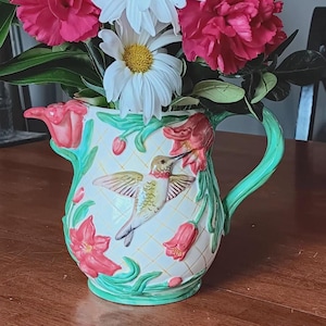 Op de afbeelding: Een decoratieve kan met een kolibrie-ontwerp, gevuld met een boeket witte madeliefjes en levendige roze anjers. De kan heeft een groen handvat en bloemaccenten, wat een charmante presentatie creëert.