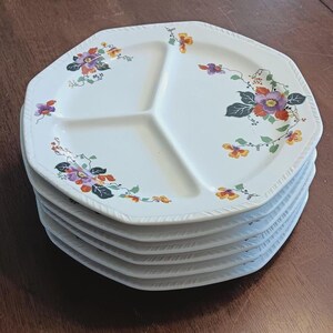 May include: A stack of six white ceramic divided plates with a floral design in shades of purple, orange, and yellow. The plates have a scalloped edge and a raised rim.