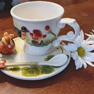 May include: A white ceramic teacup and saucer set with a duck and floral design. The set includes a spoon and a small duck figurine. White daisies and a butterfly spoon rest are arranged around the set.