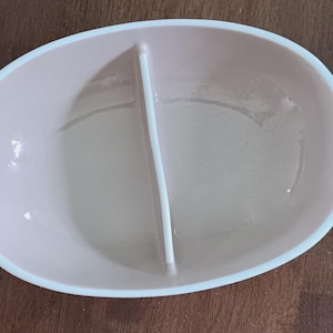 May include: A light pink, oval-shaped divided serving dish with a white rim and a central divider. The dish is empty and sits on a dark brown wooden surface. The dish is designed to hold two separate food items.