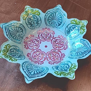 May include: A decorative glass bowl with a floral design. The bowl features a light blue exterior with teal and green accents. The center of the bowl has a pink and gold floral pattern. The bowl is on a wooden surface.