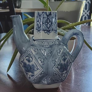 以下が含まれることがあります： 青と白の陶器のティーポットと、それに合うカップ。ティーポットは丸みを帯びたボディ、曲がった注ぎ口、ハンドルが付いています。カップは長方形で、ティーポットの上にあります。どちらの作品も花柄と幾何学模様で装飾されています。