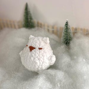 May include: A white crocheted owl plush toy with brown eyes, sitting on a white fluffy surface.