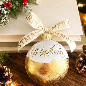 K&ouml;nnte beinhalten: Eine kugelf&ouml;rmige Weihnachtsbaumkugel mit einem wei&szlig;en oberen Bereich, auf dem der Name Madison in goldener Schreibschrift steht, und einem unteren Bereich aus strukturierter Blattgoldoptik. Ein cremefarbenes Band mit goldenem Merry Christmas-Text.