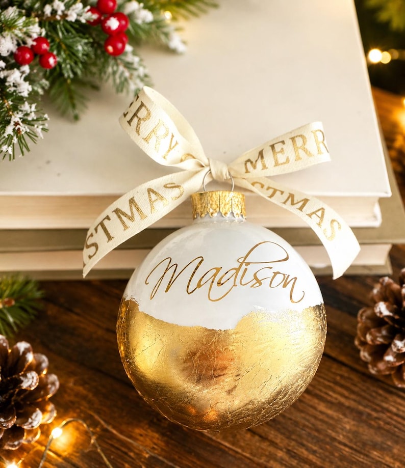 K&ouml;nnte beinhalten: Eine kugelf&ouml;rmige Weihnachtsbaumkugel mit einem wei&szlig;en oberen Bereich, auf dem der Name Madison in goldener Schreibschrift steht, und einem unteren Bereich aus strukturierter Blattgoldoptik. Ein cremefarbenes Band mit goldenem Merry Christmas-Text.