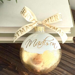 K&ouml;nnte beinhalten: Eine kugelf&ouml;rmige wei&szlig;e Weihnachtskugel mit Blattgold-Finish auf der unteren H&auml;lfte und dem Namen Madison in goldener Schreibschrift. Ein cremefarbenes Band mit dem goldenen Text Merry Christmas ist an der oberen Metallkappe befestigt.