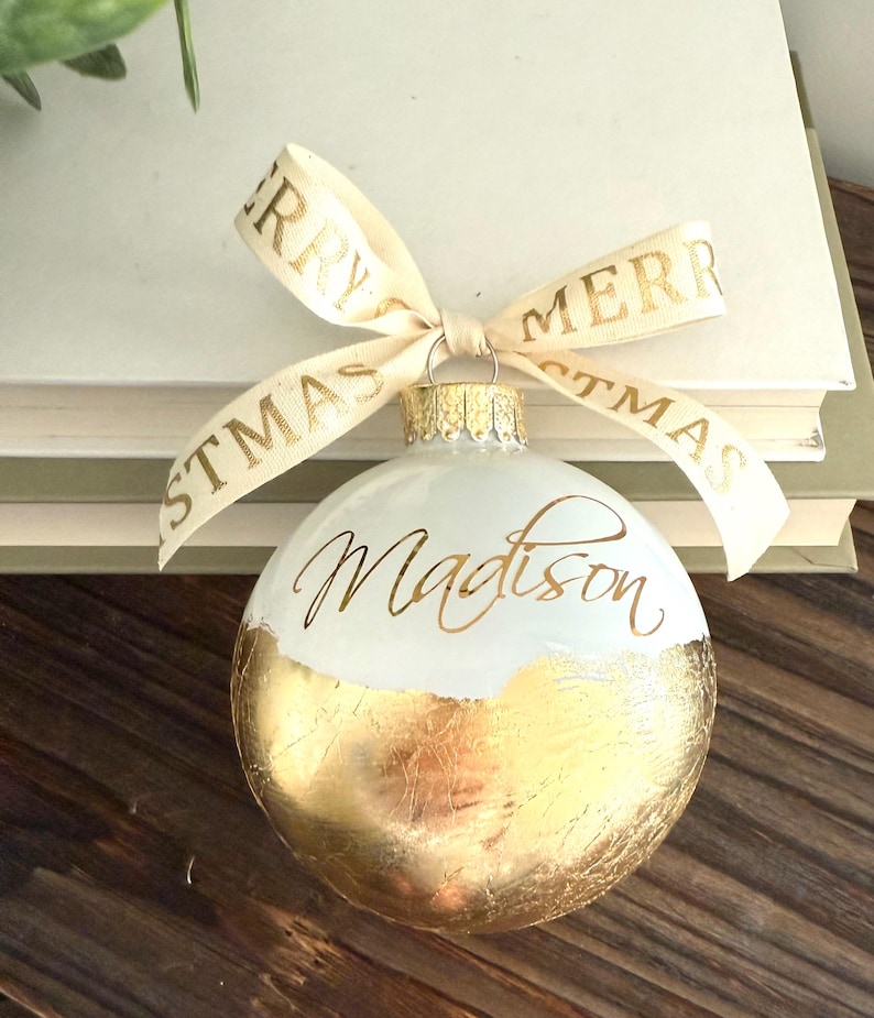 K&ouml;nnte beinhalten: Eine kugelf&ouml;rmige wei&szlig;e Weihnachtskugel mit Blattgold-Finish auf der unteren H&auml;lfte und dem Namen Madison in goldener Schreibschrift. Ein cremefarbenes Band mit dem goldenen Text Merry Christmas ist an der oberen Metallkappe befestigt.