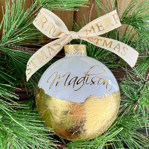 K&ouml;nnte beinhalten: Eine kugelf&ouml;rmige wei&szlig;e Weihnachtskugel mit einer Basis aus Blattgold und dem in goldener Schreibschrift verfassten Namen Madison. Ein cremefarbenes Band mit dem goldenen Schriftzug Merry Christmas ist als Schleife an der Metallkappe befestigt.