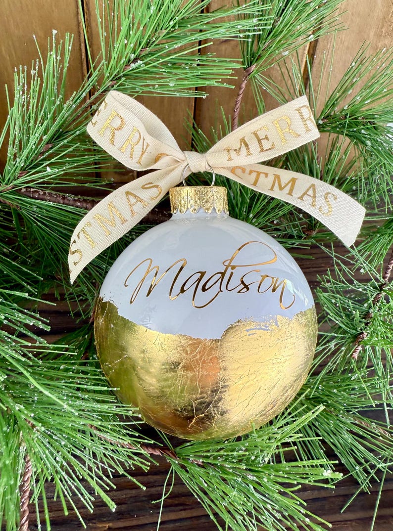 K&ouml;nnte beinhalten: Eine kugelf&ouml;rmige wei&szlig;e Weihnachtskugel mit einer Basis aus Blattgold und dem in goldener Schreibschrift verfassten Namen Madison. Ein cremefarbenes Band mit dem goldenen Schriftzug Merry Christmas ist als Schleife an der Metallkappe befestigt.