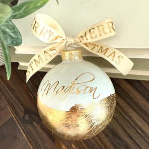 K&ouml;nnte beinhalten: Eine kugelf&ouml;rmige wei&szlig;e Weihnachtskugel mit dem Namen Madison in goldener Schreibschrift. Die untere H&auml;lfte der Kugel ist mit Blattgold bedeckt. Ein cremefarbenes Band mit dem goldenen Text Merry Christmas ist oben festgebunden.