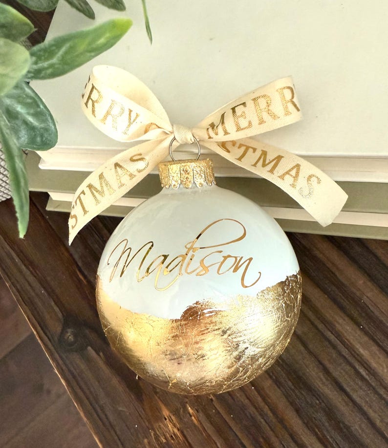 K&ouml;nnte beinhalten: Eine kugelf&ouml;rmige wei&szlig;e Weihnachtskugel mit dem Namen Madison in goldener Schreibschrift. Die untere H&auml;lfte der Kugel ist mit Blattgold bedeckt. Ein cremefarbenes Band mit dem goldenen Text Merry Christmas ist oben festgebunden.