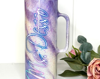 May include: Purple and white glitter tumbler with a handle and a lid. The tumbler has the name "Miss Dess" written on it in a blue and pink script font.