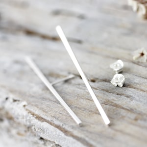 May include: A pair of silver bar earrings with a minimalist design. The earrings are long and thin, and they have a simple, sleek look.