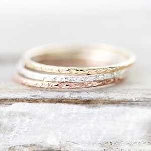 May include: Three thin gold rings stacked on top of each other. The top ring is yellow gold, the middle ring is silver, and the bottom ring is rose gold. All three rings have a hammered texture.