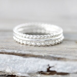 May include: Three silver stacking rings. The bottom ring has a beaded design, the middle ring is a plain band, and the top ring has a twisted rope design.
