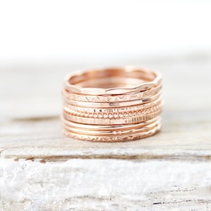 Peut inclure: Une pile de bagues empilables en or rose avec différentes textures et motifs. Les bagues sont disposées en cercle et se trouvent sur une surface en bois blanc.