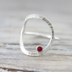 May include: A silver ring with a textured, circular band and a small, red gemstone set in a gold bezel.