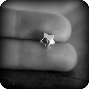 May include: A small, shiny, silver star-shaped charm. The charm is in sharp focus, with the details of the star's points and surface clearly visible. The charm is held between two fingers, with a blurred background.