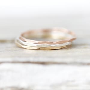 May include: Three thin hammered metal rings stacked together. The top ring is rose gold, the middle ring is silver, and the bottom ring is gold.