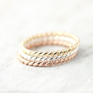May include: Three thin gold rings, one in rose gold, one in yellow gold, and one in silver, are stacked on top of each other. The rings are all twisted or rope-style.