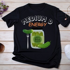 Puede incluir: Camiseta negra con el texto "Medium D Energy" en blanco y naranja. Se muestra un frasco de pepinillos encurtidos con un pepinillo en un tenedor.
