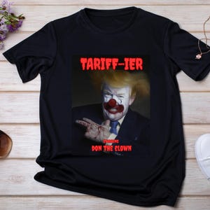 Könnte beinhalten: Schwarzes T-Shirt mit einer Grafik einer als Clown geschminkten Person, mit dem Text "TARIFF-IER" darüber und "DON the CLOWN" darunter. Das Bild befindet sich auf einer Holzoberfläche mit Zubehör.