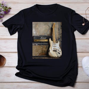 May include: Black t-shirt with a graphic print of a vintage electric guitar and amplifier. The guitar is white with a worn finish and has a brown pickguard. The amplifier is black with a white logo that says "Marshall".