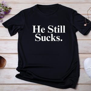 May include: Black t-shirt with the white text "He Still Sucks." The shirt is displayed on a light wood surface, with sunglasses, white sneakers, and purple flowers nearby. A bottle of nail polish and a necklace are also visible.