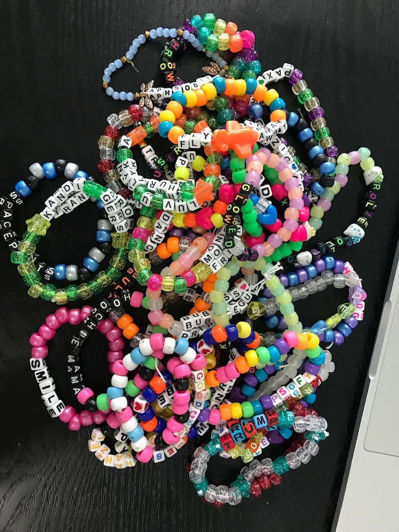 May include: A pile of colorful beaded bracelets with words spelled out on them, including "Smile", "Good", "Trump", "Space", "Kiran", "Mama", "Fly", "P.S.O.F", "Good", "Moon", "Gloe", "Rollin", "Bub", "Egu", "Hur", "Luv", "Dance", "Aphrodite", "100", "Pax", "Arrow", and more.