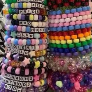 May include: A collection of colorful beaded bracelets with various words and designs, including "Pride", "Moo", "Plur", "Violent", "Apollo", "Nihachu", "My Godz", and "Boo".