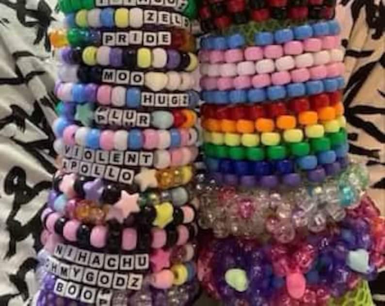 May include: A collection of colorful beaded bracelets with various words and designs, including "Pride", "Moo", "Plur", "Violent", "Apollo", "Nihachu", "My Godz", and "Boo".