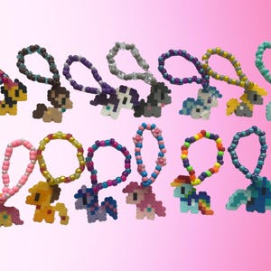 May include: A collection of beaded pony keychains in various colors. Each keychain features a pony design made from colorful beads, with a loop of beads for attachment. The ponies are in different colors and styles, with a pink background.