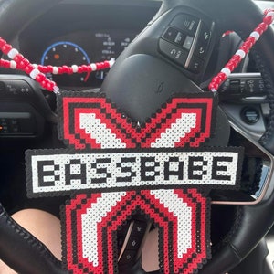 Puede incluir: Un amuleto de coche de cuentas rojas, blancas y negras con la palabra "BEESEEBEE" en cuentas blancas. El amuleto está colgado del volante de un coche.