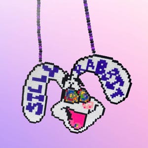 May include: A pixelated bunny pendant necklace with a white body, purple accents, and a colorful eye design. The bunny's ears display the text "STILL FAB". The necklace chain is made of purple and black beads.