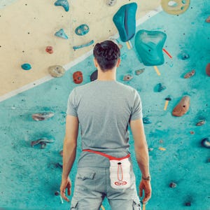 May include: A person stands before a climbing wall, wearing a gray t-shirt and cargo pants. A white and red climbing chalk bag with the word "Yogurt" is attached to the waist with a red belt. The climbing wall has various colored handholds.