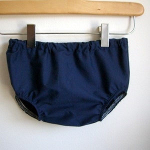 May include: A navy blue baby bloomer with a white elastic waistband and trim. The bloomer is hanging on a wooden hanger.