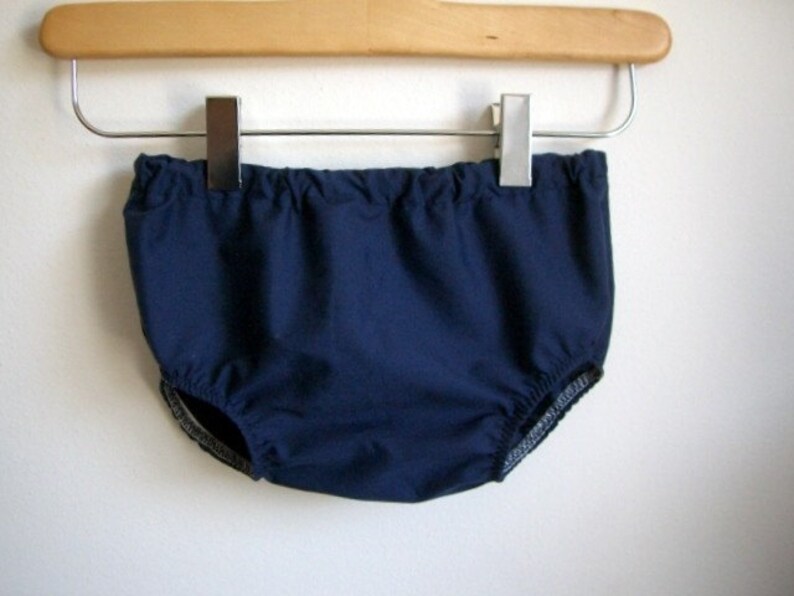 May include: A navy blue baby bloomer with a white elastic waistband and trim. The bloomer is hanging on a wooden hanger.