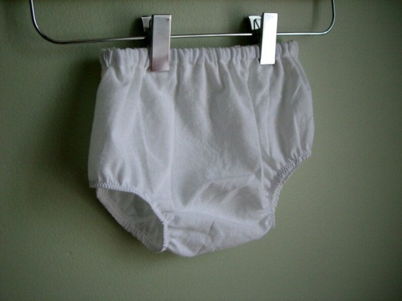 May include: White cotton baby bloomers hanging on a clothes hanger.